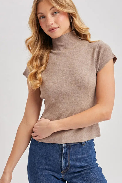 A woman wearing a taupe-brown short-sleeve mock neck knit top and blue jeans, photographed against a light gray studio background.