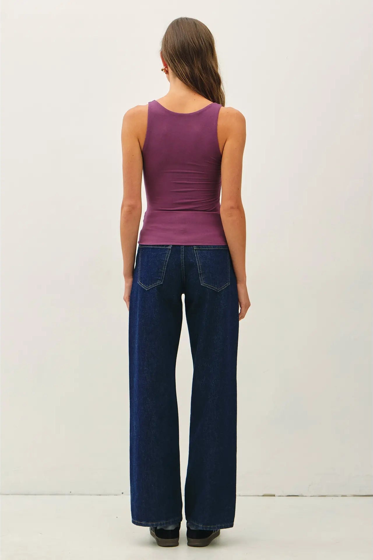 A woman viewed from the back wearing a sleeveless purple tank top and dark blue wide-leg jeans against a plain off-white studio background.