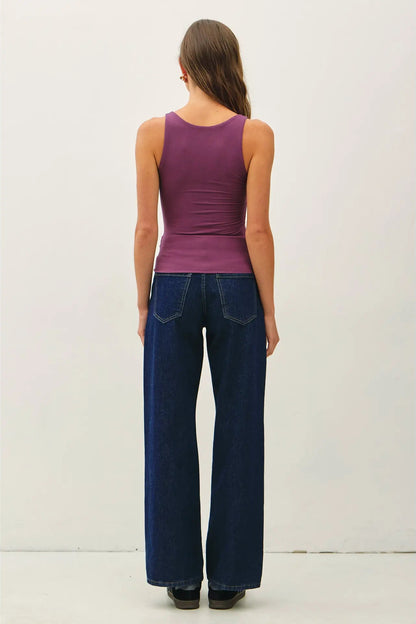A woman viewed from the back wearing a sleeveless purple tank top and dark blue wide-leg jeans against a plain off-white studio background.