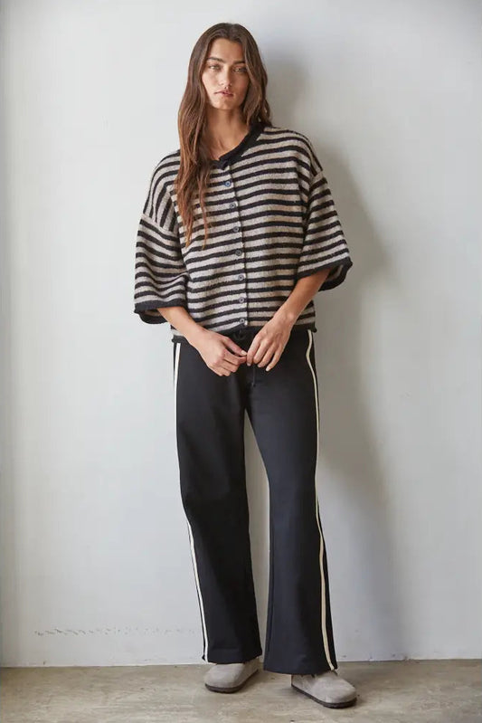 A woman stands against a plain light gray wall wearing a loose black-and-white horizontally striped zip-front top with wide three-quarter sleeves, black wide-leg pants with white side stripes, and light gray slip-on shoes.