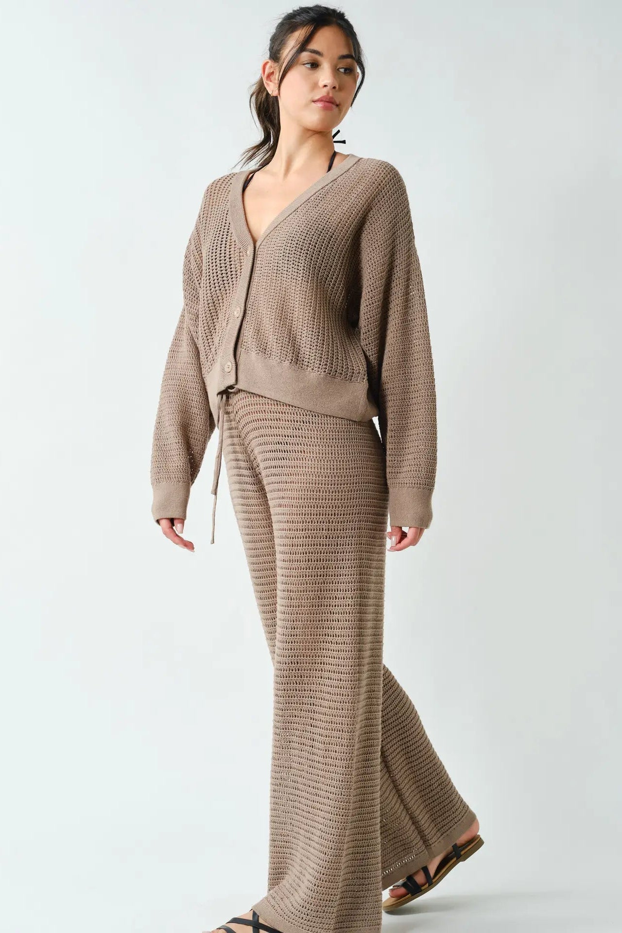 A woman modeling a matching knit lounge set consisting of a cropped cardigan and wide-leg pants in a taupe brown color, set against a plain light gray background.