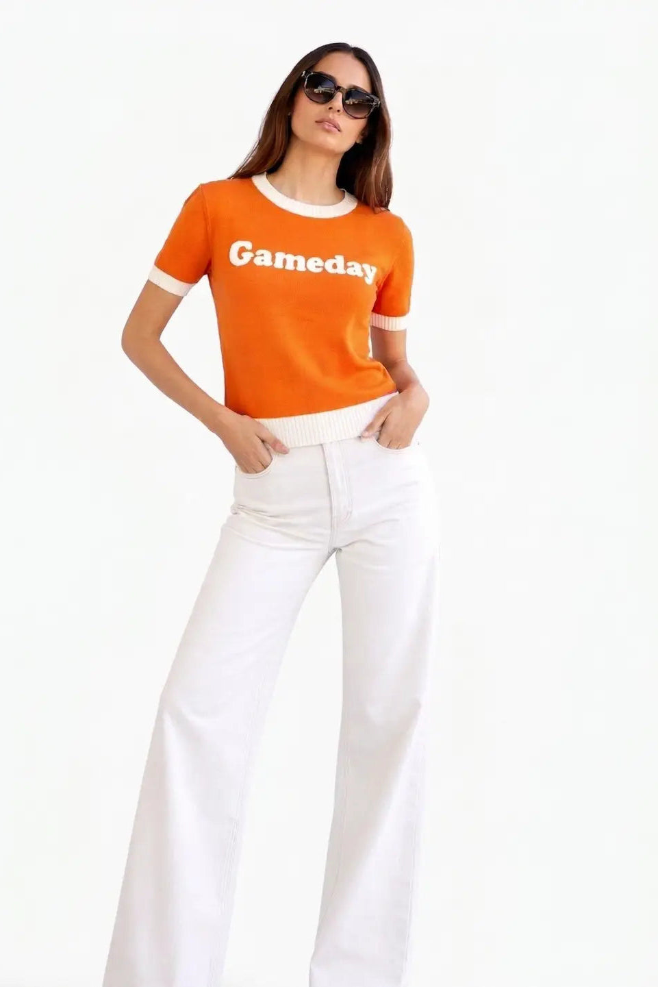 A woman modeling a bright orange short-sleeve ringer T-shirt with white trim and the word 'Gameday' printed across the chest, paired with white pants, against a plain white background.