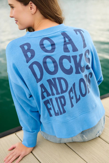 Person wearing a blue sweatshirt with 'BOAT DOCKS AND FLIP FLOPS' text by a body of water.