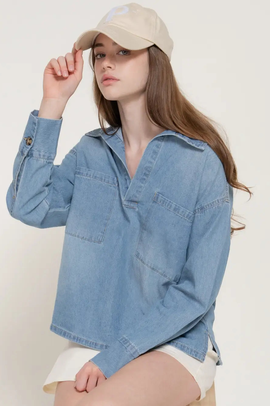 A woman wearing a light blue denim long-sleeve shirt with a relaxed fit, paired with a beige baseball cap and white shorts, posing against a plain off-white background.
