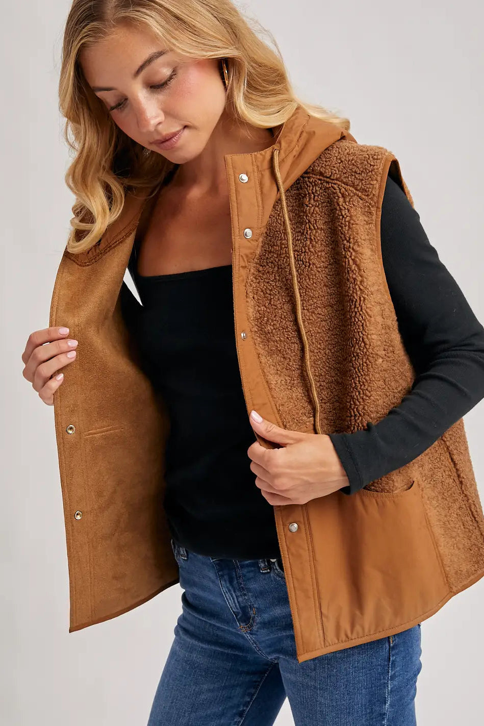 A woman wearing a brown sherpa-lined suede hooded vest over a black long-sleeve top and blue jeans, shown against a light gray studio background.