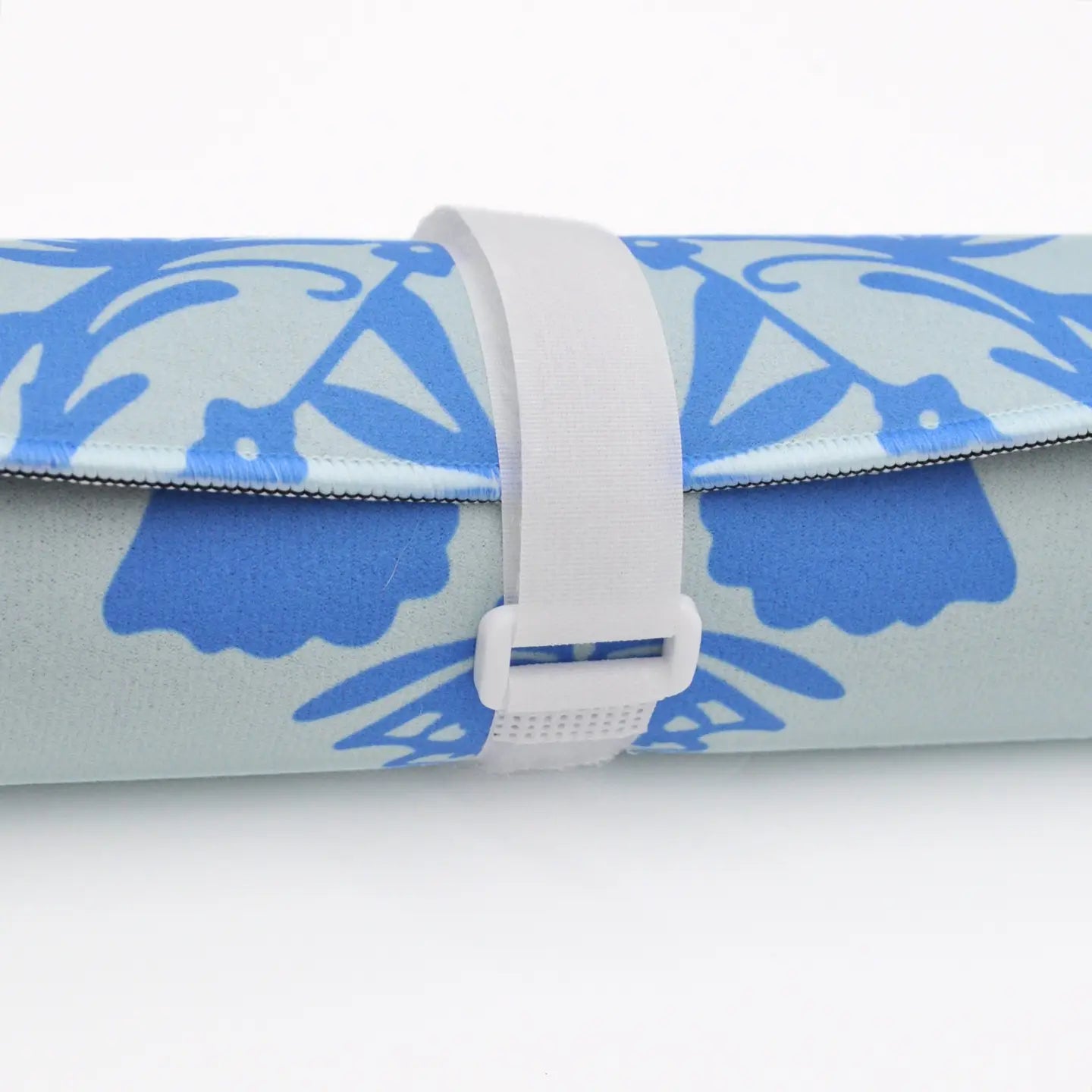 A rolled-up yoga mat with a light blue and medium blue floral pattern, secured with a white elastic strap and plastic buckle, on a white background.