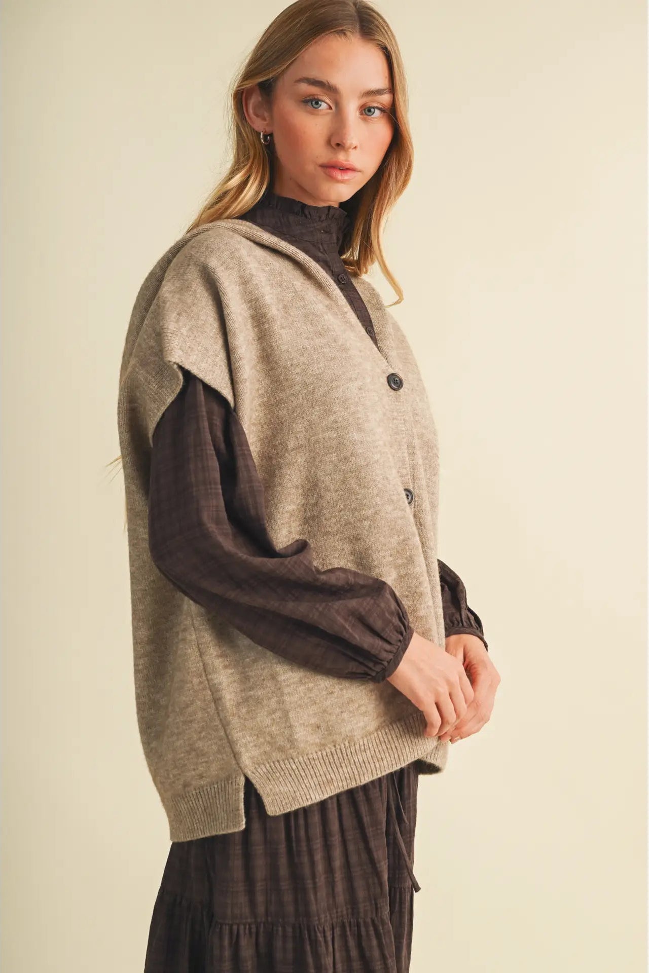 A woman modeling a sleeveless taupe knit vest layered over a dark brown long-sleeve top and a dark brown pleated skirt, set against a plain beige studio background.