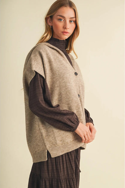 A woman modeling a sleeveless taupe knit vest layered over a dark brown long-sleeve top and a dark brown pleated skirt, set against a plain beige studio background.