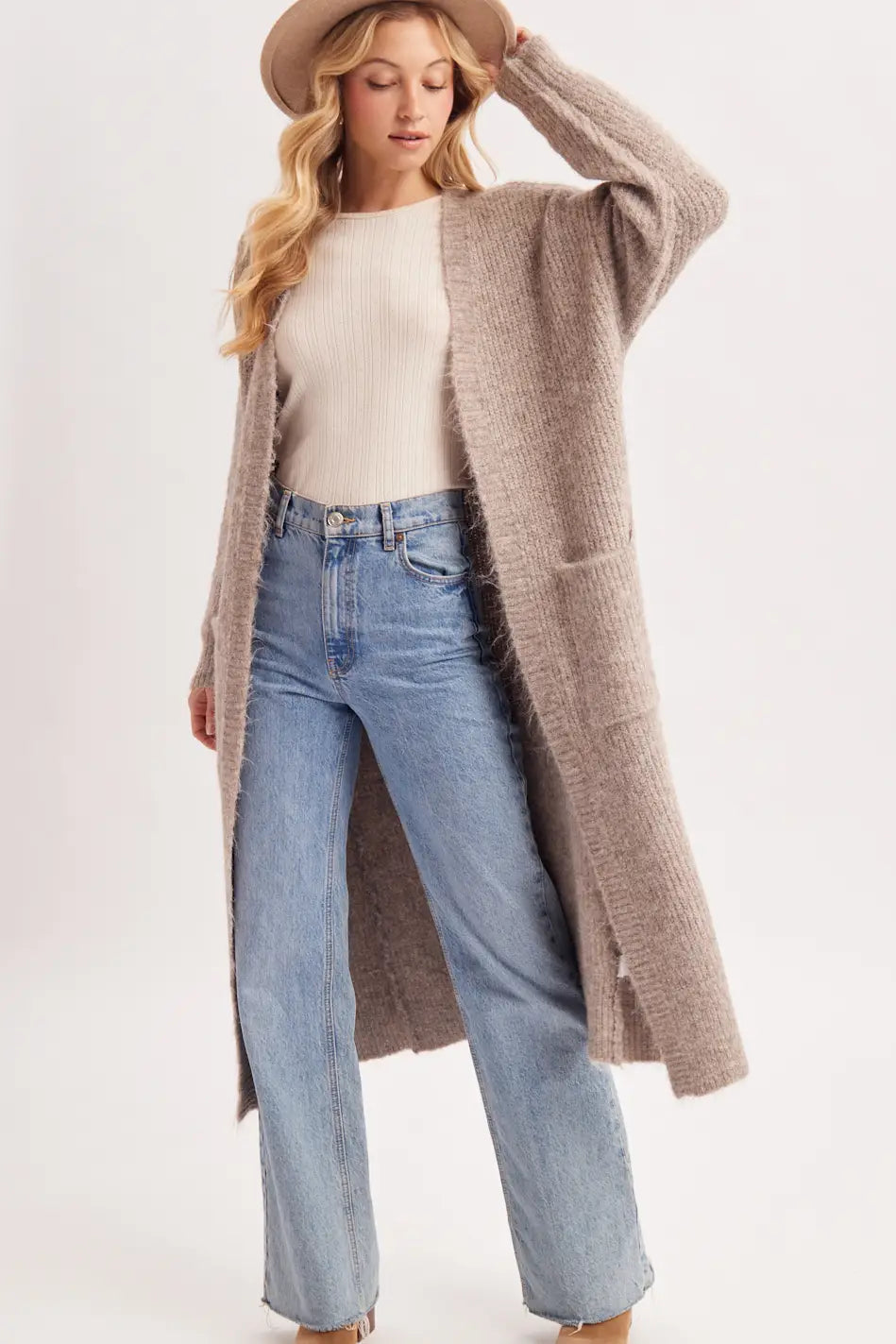 A woman modeling a long, open-front chunky knit cardigan in taupe-beige, worn over a cream ribbed top and light-wash blue jeans, with a tan wide-brim hat against a plain light background.
