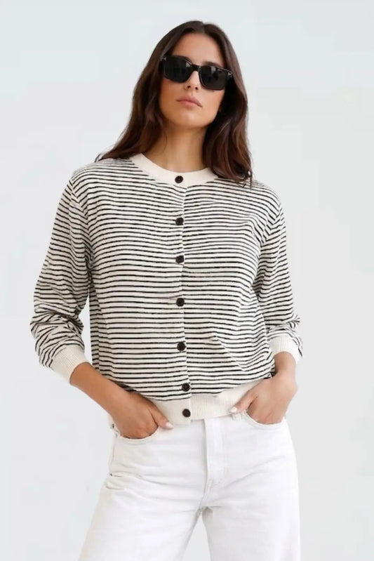 A woman wearing a black and white horizontally striped button-up cardigan sweater with white pants and black sunglasses, standing against a plain light gray background.