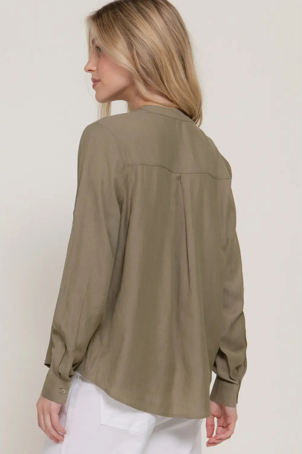 A woman shown from the back wearing a loose, long-sleeve button-up blouse in a muted olive-taupe color, paired with white pants, against a plain light beige studio background.
