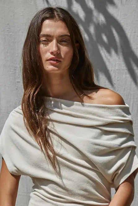 A woman with long brown hair wearing a draped, off-the-shoulder cream-colored top, standing against a textured gray wall with leaf shadows.