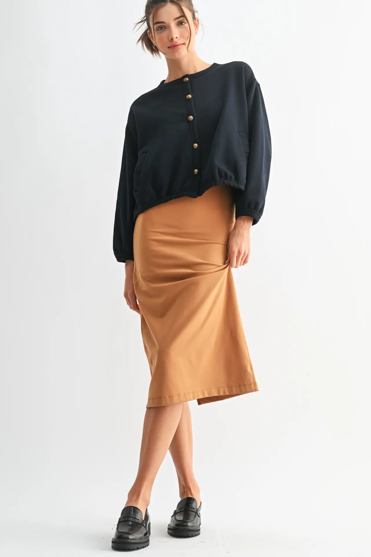 A woman modeling a black long-sleeve button-front top and a tan midi skirt, paired with black loafers, against a plain light gray studio background.