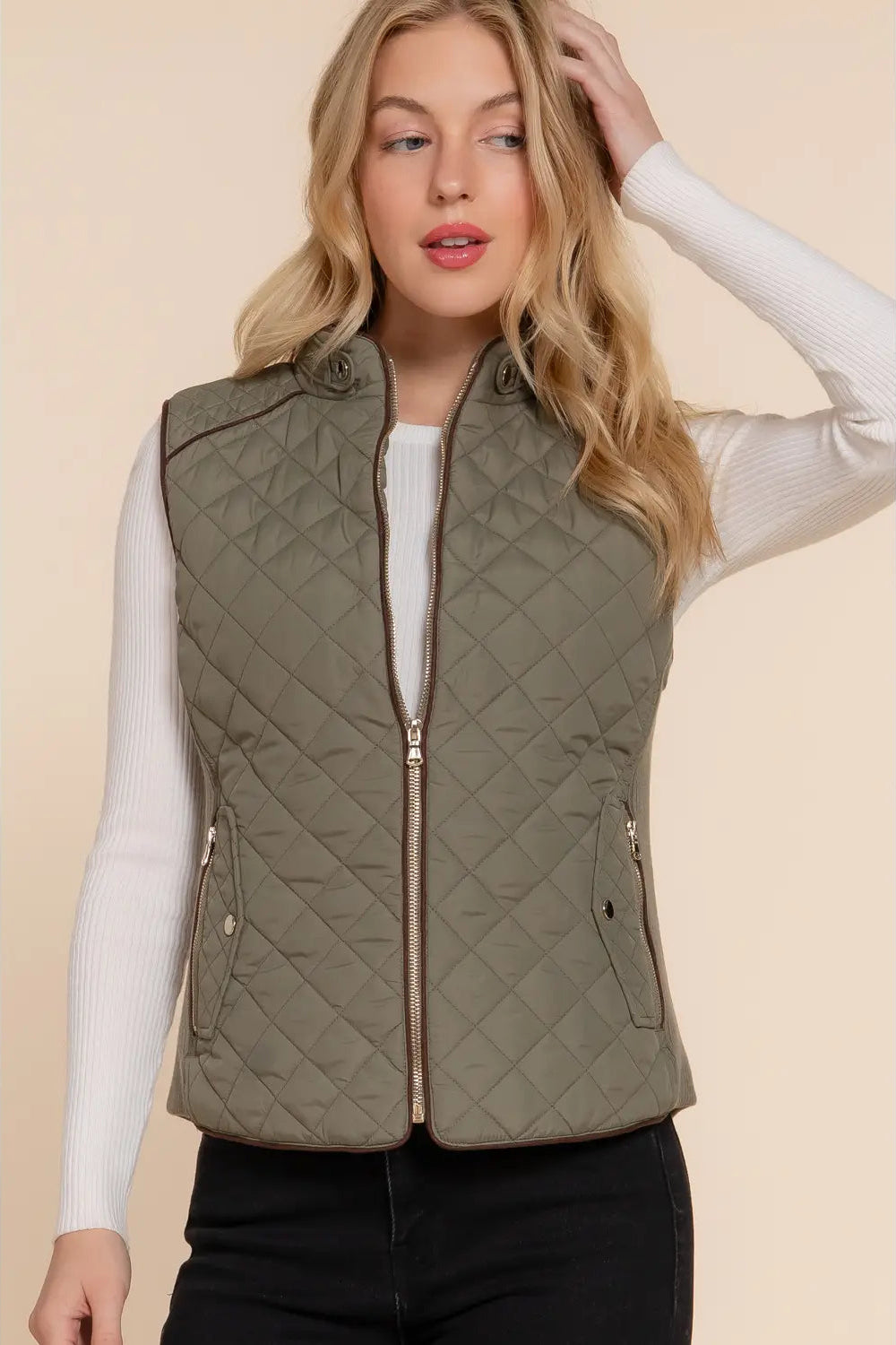 A woman wearing an olive green diamond-quilted zip-front vest over a white long-sleeve top and black pants, posed against a solid beige background.