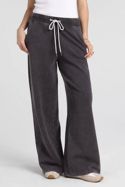 A person wearing dark charcoal wide-leg sweatpants with a white drawstring, paired with a white top and beige sneakers, shown against a light gray studio background.