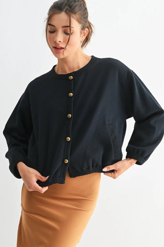 A woman wearing a black cropped button-front jacket with gold-tone buttons and a tan skirt, photographed against a plain light background.