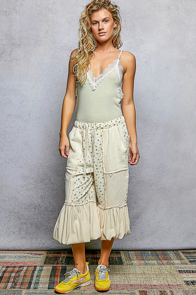 A woman modeling a light sage green camisole top with white lace trim and a matching cream patchwork midi skirt with embroidered details, paired with yellow sneakers, standing against a textured gray wall.