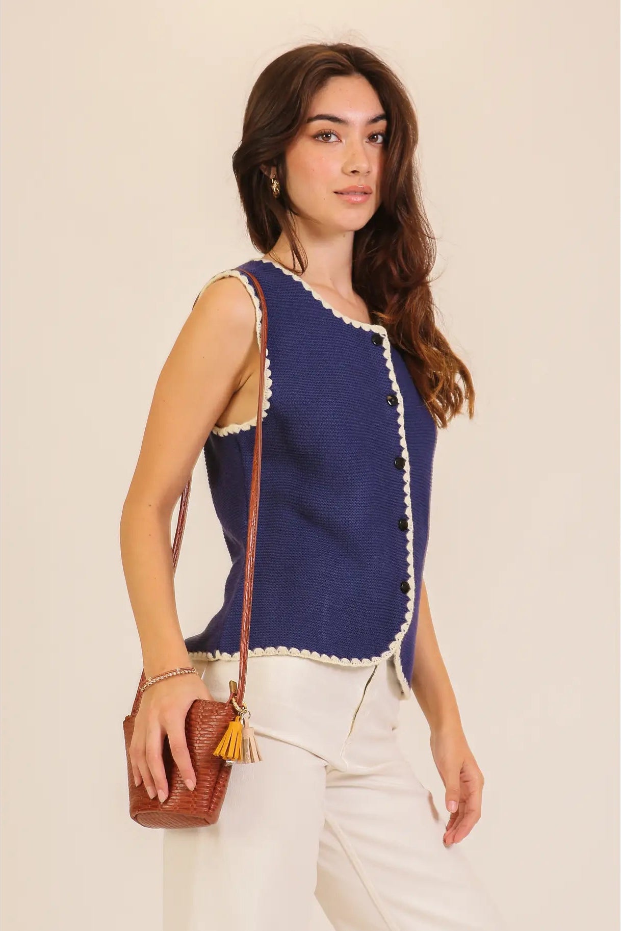 A woman modeling a sleeveless navy blue knit vest with cream scalloped trim and front buttons, paired with white pants and a small brown crossbody bag, against a plain beige studio background.