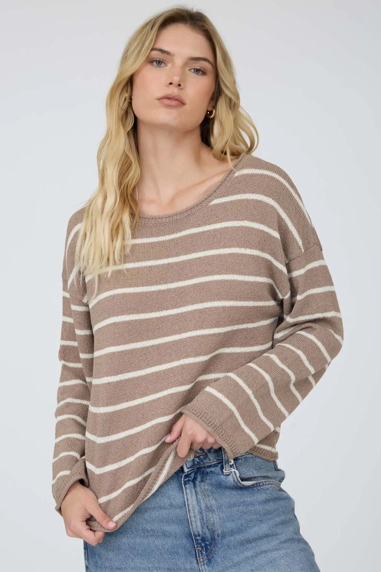 A woman wearing a brown and cream striped knit sweater with blue jeans, photographed against a plain light gray studio background.