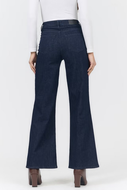 Rear view of a person wearing dark blue wide-leg flare jeans with a white long-sleeve top and brown heeled shoes against a light gray studio background.