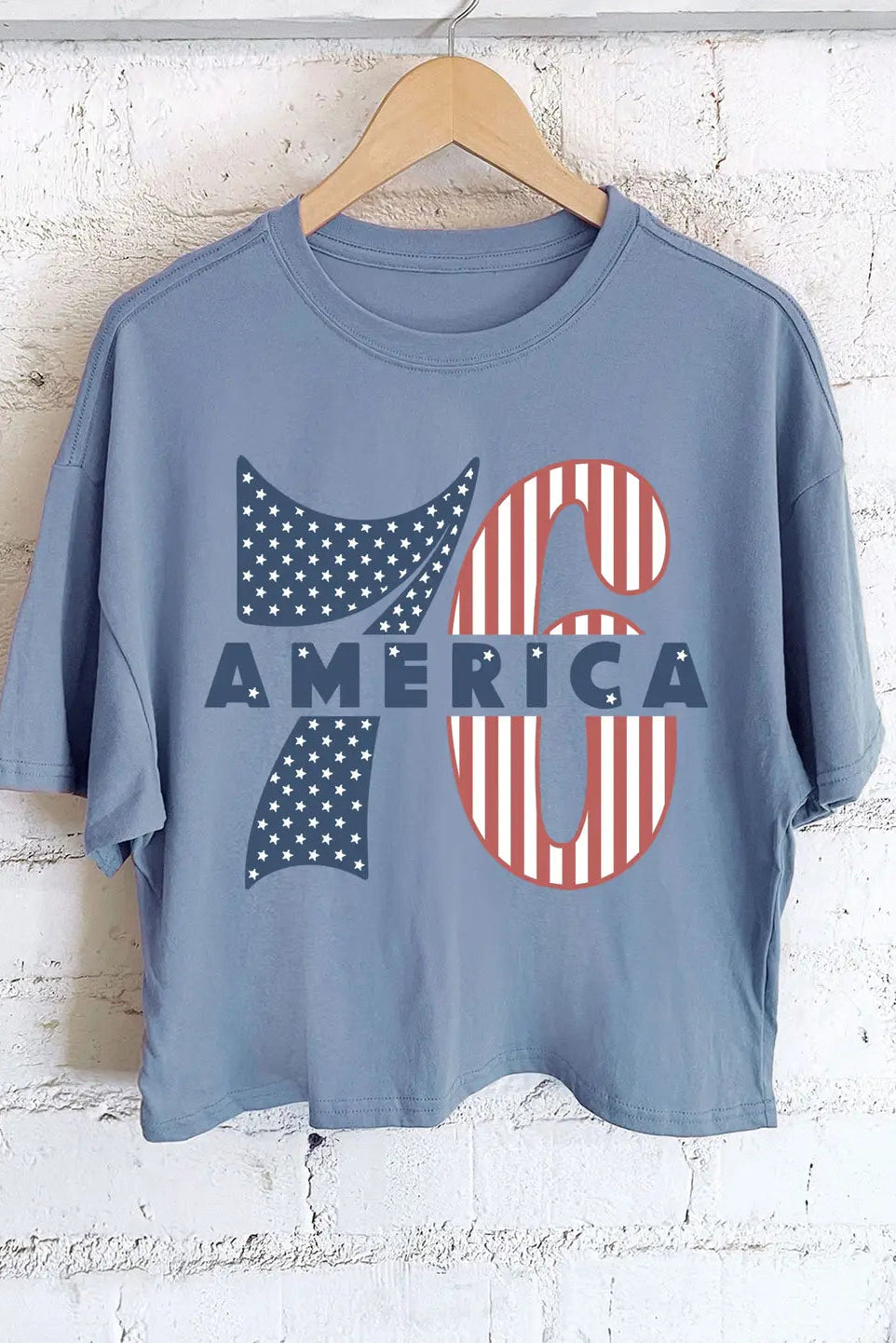Blue t-shirt with '76 America' design on a wooden hanger against a white brick wall.