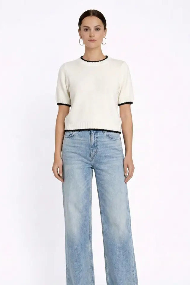 A woman modeling a short-sleeve cream knit top with black trim at the collar and sleeve cuffs, paired with light-wash wide-leg jeans, against a plain white studio background.