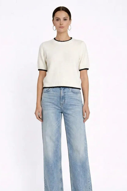 A woman modeling a short-sleeve cream knit top with black trim at the collar and sleeve cuffs, paired with light-wash wide-leg jeans, against a plain white studio background.