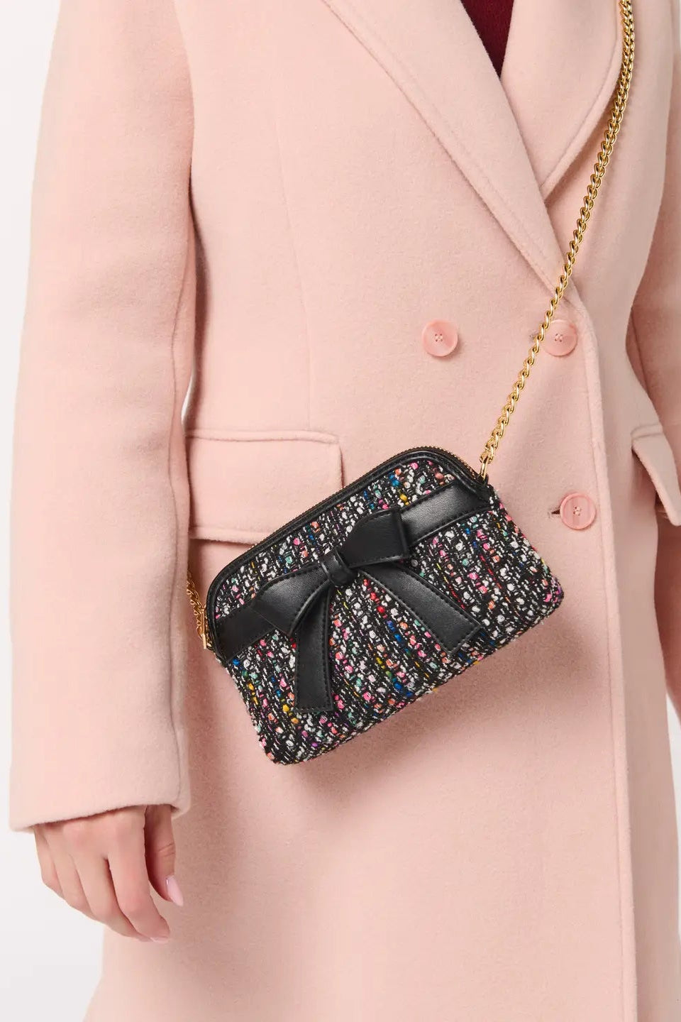 A model wearing a light pink double-breasted coat carries a small tweed crossbody bag with a black bow and gold chain strap.