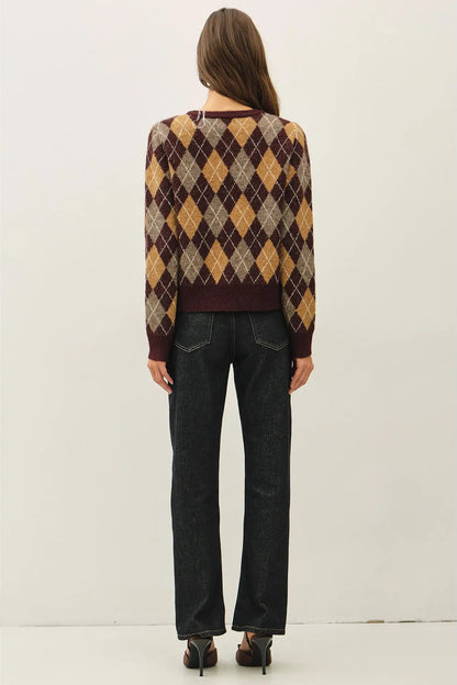 A woman viewed from the back wearing a long-sleeve argyle sweater with dark wash jeans and heels, standing against a plain light gray studio background.