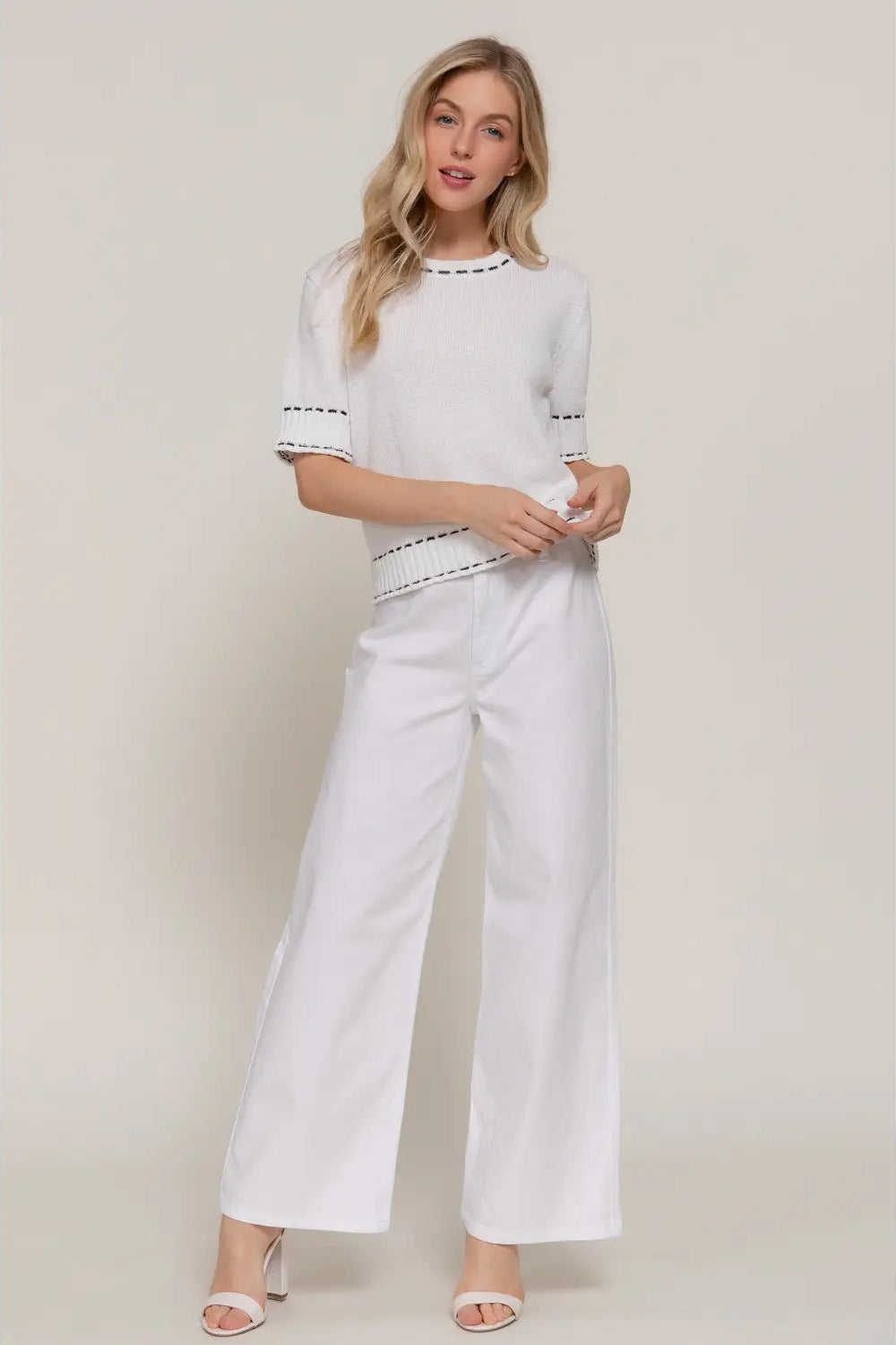 A woman modeling a white short-sleeve top with black embroidered trim and matching white wide-leg pants against a plain beige studio background.