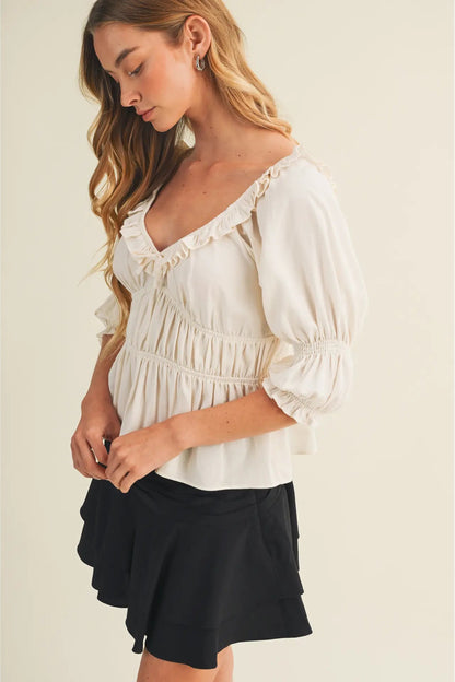 A woman wearing a cream puff-sleeve blouse with ruffle trim and a black tiered skirt, standing against a plain light beige background.