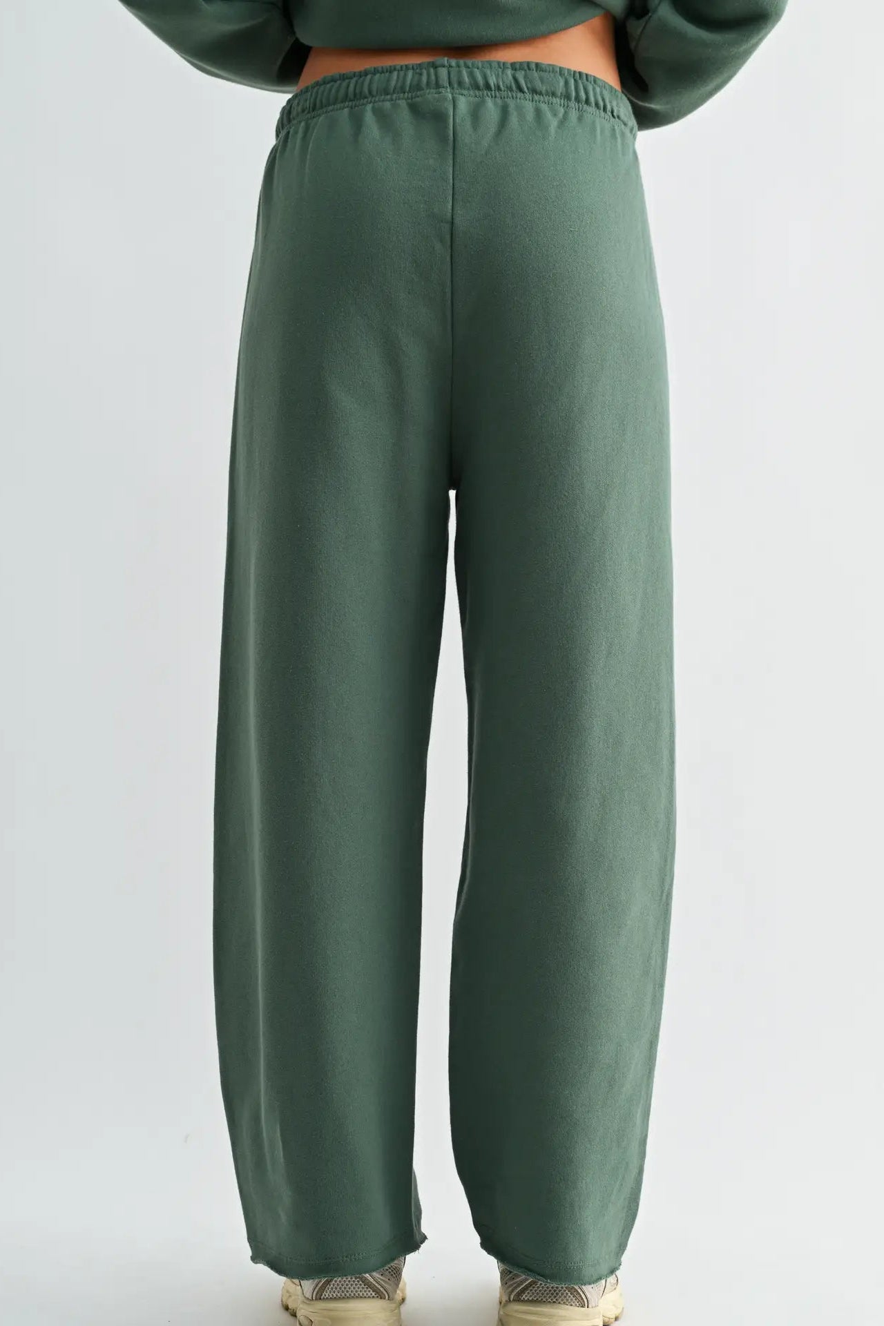 A person shown from the back wearing loose, wide-leg sage green sweatpants with an elastic waistband and cuffs, paired with a matching green cropped top and beige sneakers, against a light gray studio background.