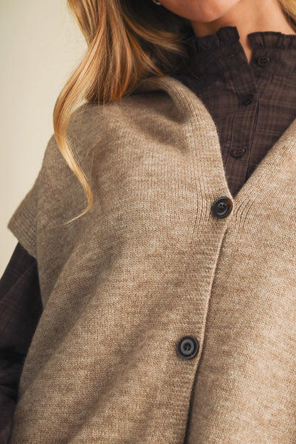 Close-up of a person wearing a taupe knit cardigan vest over a dark brown plaid button-up shirt. The cardigan has a soft, textured knit and dark buttons.