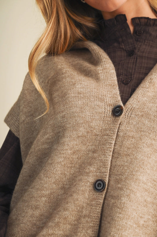 Close-up of a person wearing a taupe knit cardigan vest over a dark brown plaid button-up shirt. The cardigan has a soft, textured knit and dark buttons.