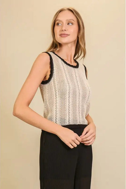 A woman modeling a sleeveless cream knit top with black trim, paired with black pants, against a solid beige background.