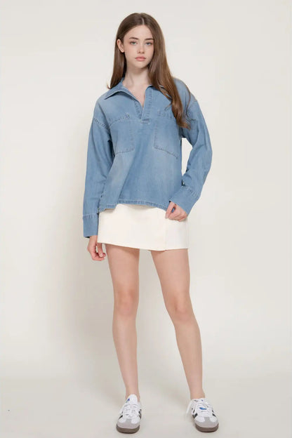 A woman modeling a light blue denim long-sleeve shirt with a half-placket and chest pockets, paired with a white mini skirt and white sneakers, against a plain light beige studio background.