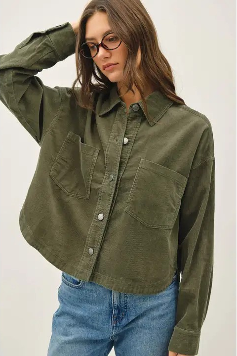 A woman wearing an olive green cropped button-up shirt with two chest pockets and blue jeans, photographed against a plain light background.