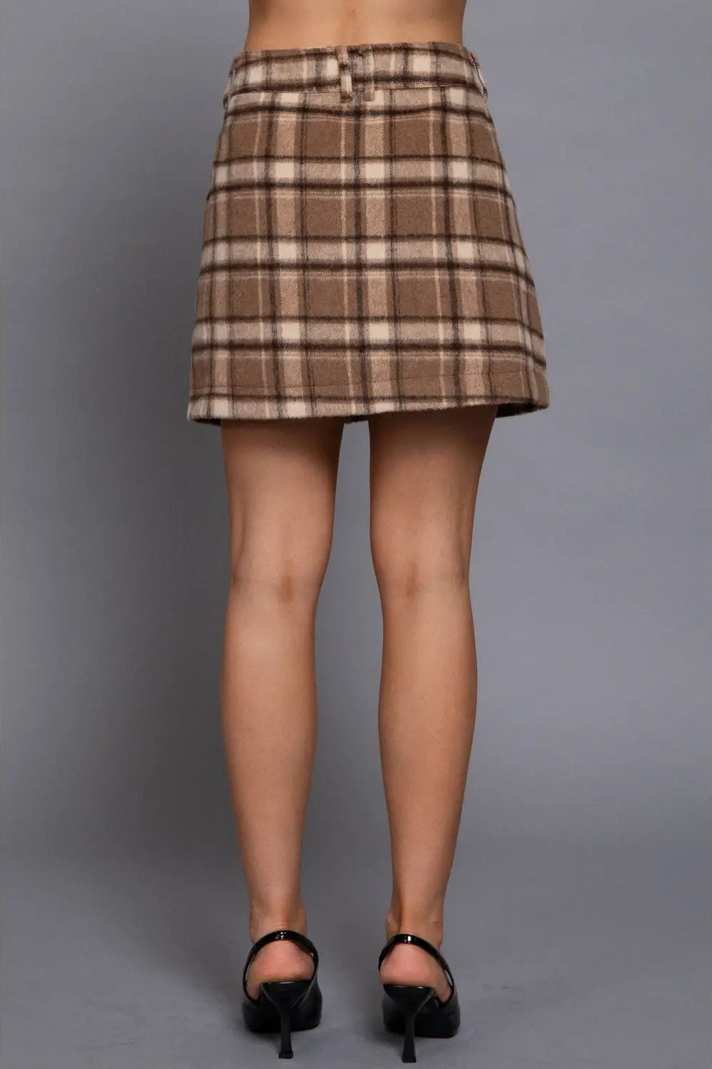 A model's lower body wearing a brown and beige plaid mini skirt with a matching belt, paired with black high heels, against a solid gray studio background.
