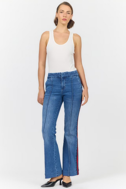 A model wearing blue denim flare jeans with red and black side stripes, paired with a white tank top and black heels, against a light gray studio background.