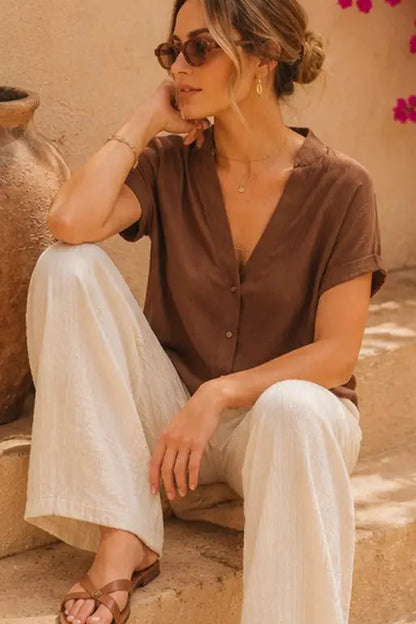 A woman wearing a brown short-sleeve button-front blouse and white wide-leg pants sits on stone steps outdoors next to a large clay pot.