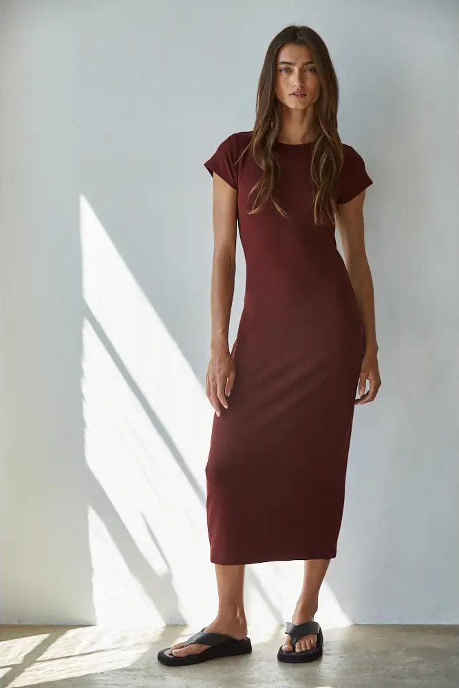 A woman modeling a short-sleeve, knee-length burgundy knit dress with black slide sandals, standing in a brightly lit room with diagonal sunlight shadows on a white wall.
