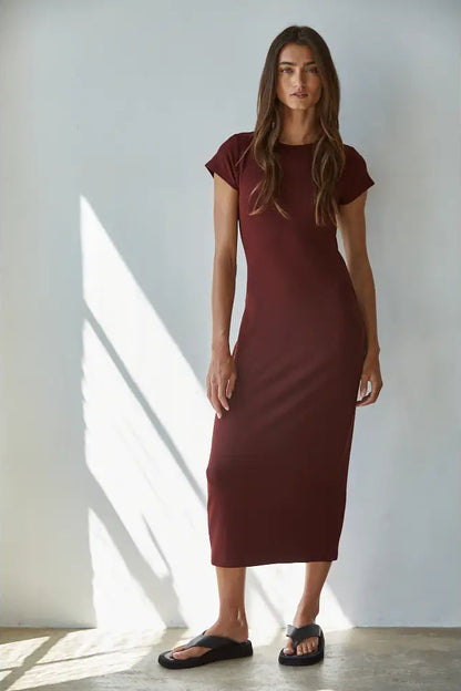 A woman modeling a short-sleeve, knee-length burgundy knit dress with black slide sandals, standing in a brightly lit room with diagonal sunlight shadows on a white wall.