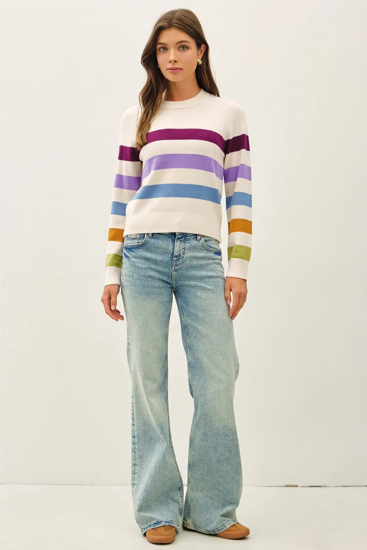 A woman modeling a white crewneck sweater with horizontal stripes in purple, blue, green, orange, and yellow, paired with light-wash flare jeans and tan shoes, against a plain off-white studio background.