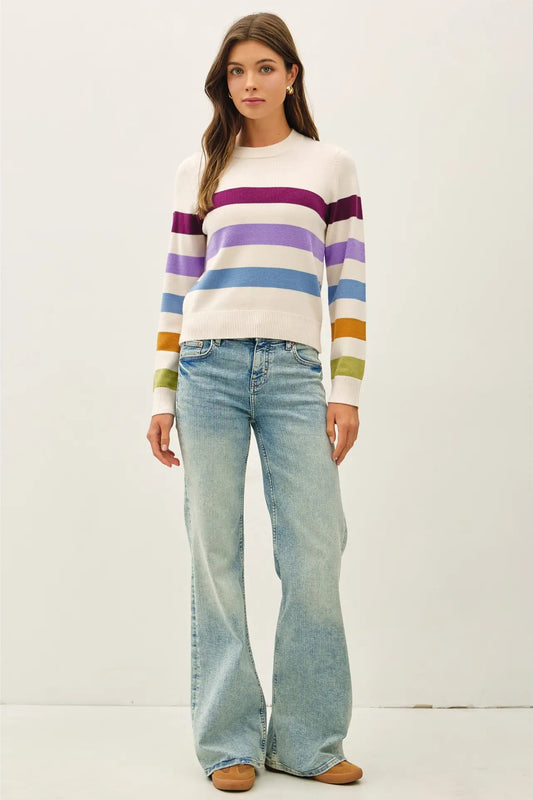 A woman modeling a white crewneck sweater with horizontal stripes in purple, blue, green, orange, and yellow, paired with light-wash flare jeans and tan shoes, against a plain off-white studio background.