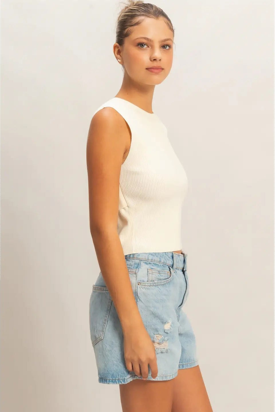 Woman wearing a white sleeveless top and denim shorts on a plain background