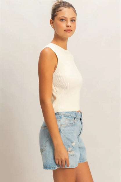 Woman wearing a white sleeveless top and denim shorts on a plain background