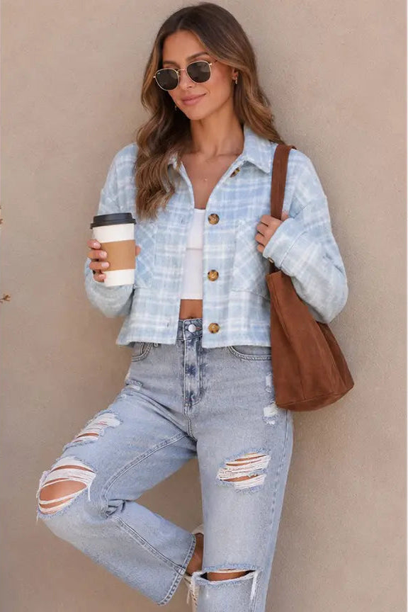 A woman wearing a light blue and white plaid cropped button-up shirt, distressed light-wash jeans, sunglasses, and carrying a brown shoulder bag, holding a coffee cup against a beige wall.