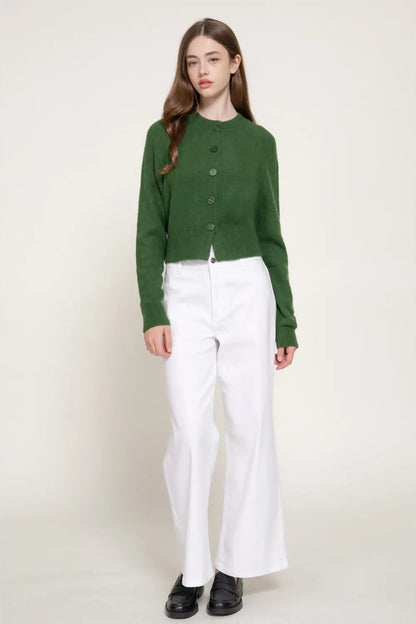 A model wearing a green cropped button-up knit cardigan and white wide-leg trousers, standing against a plain light beige studio background.
