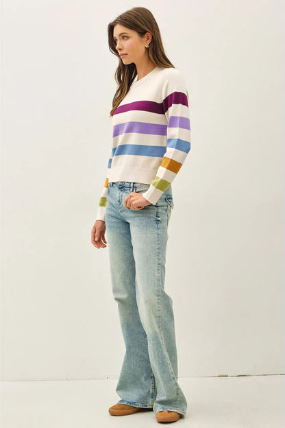 A woman modeling a white long-sleeve knit sweater with horizontal stripes in purple, blue, green, and yellow, paired with light-wash flare jeans and tan shoes, against a plain white studio background.