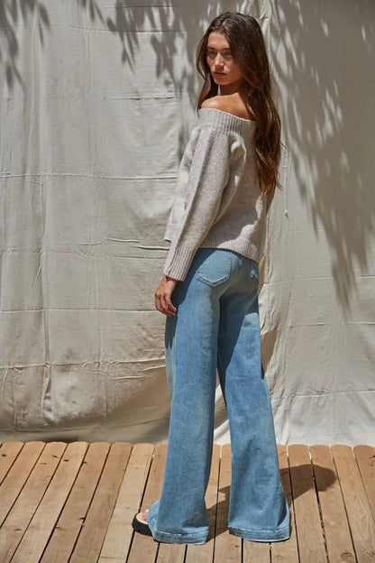 A woman stands on a wooden deck in front of a draped fabric backdrop, wearing a light gray off-the-shoulder knit sweater and light blue wide-leg jeans.